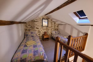 Ein kleiner, gemütlicher Raum mit schrägem Dach, zwei Einzelbetten mit gemusterten Betttextilien, einem kleinen Holfnachtkästchen und einem gerahmten Bild an der Wand.