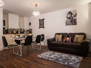 A modern living room with a dark brown sofa, a dining table with chairs, and a kitchen in the background. - Sav Rutland Town Centre-3 Bed Apartment (Leicester)