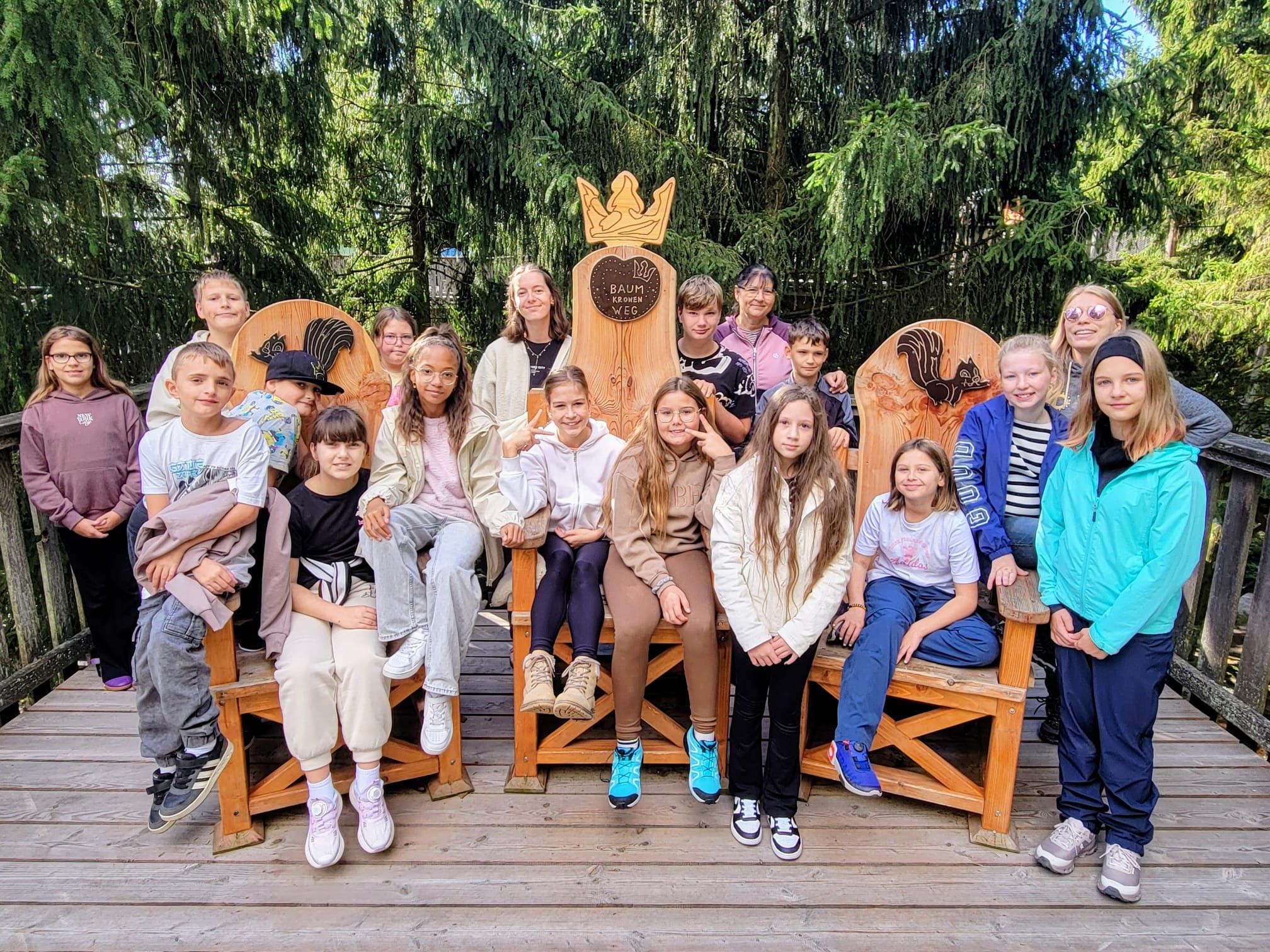 A group of people seated on wooden benches outdoors with a wooden structure featuring a crown design in the background.