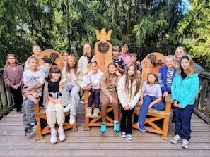 A group of people seated on wooden benches outdoors with a wooden structure featuring a crown design in the background. - Baumkronenweg GmbH (Kopfing im Innkreis)