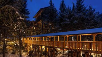 A wooden deck with a glass enclosure, surrounded by snow-covered trees and a building structure in the background.