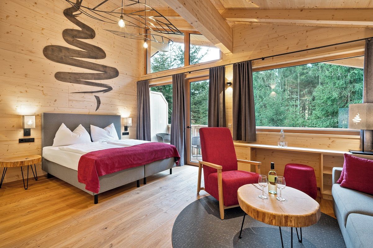 Ein modernes Hotelzimmer mit einem Bett, einem roten Sessel, einem runden Holztisch und einem großen Fenster mit Blick auf den Wald.