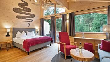 Ein modernes Hotelzimmer mit einem Bett, einem roten Sessel, einem runden Holztisch und einem großen Fenster mit Blick auf den Wald.