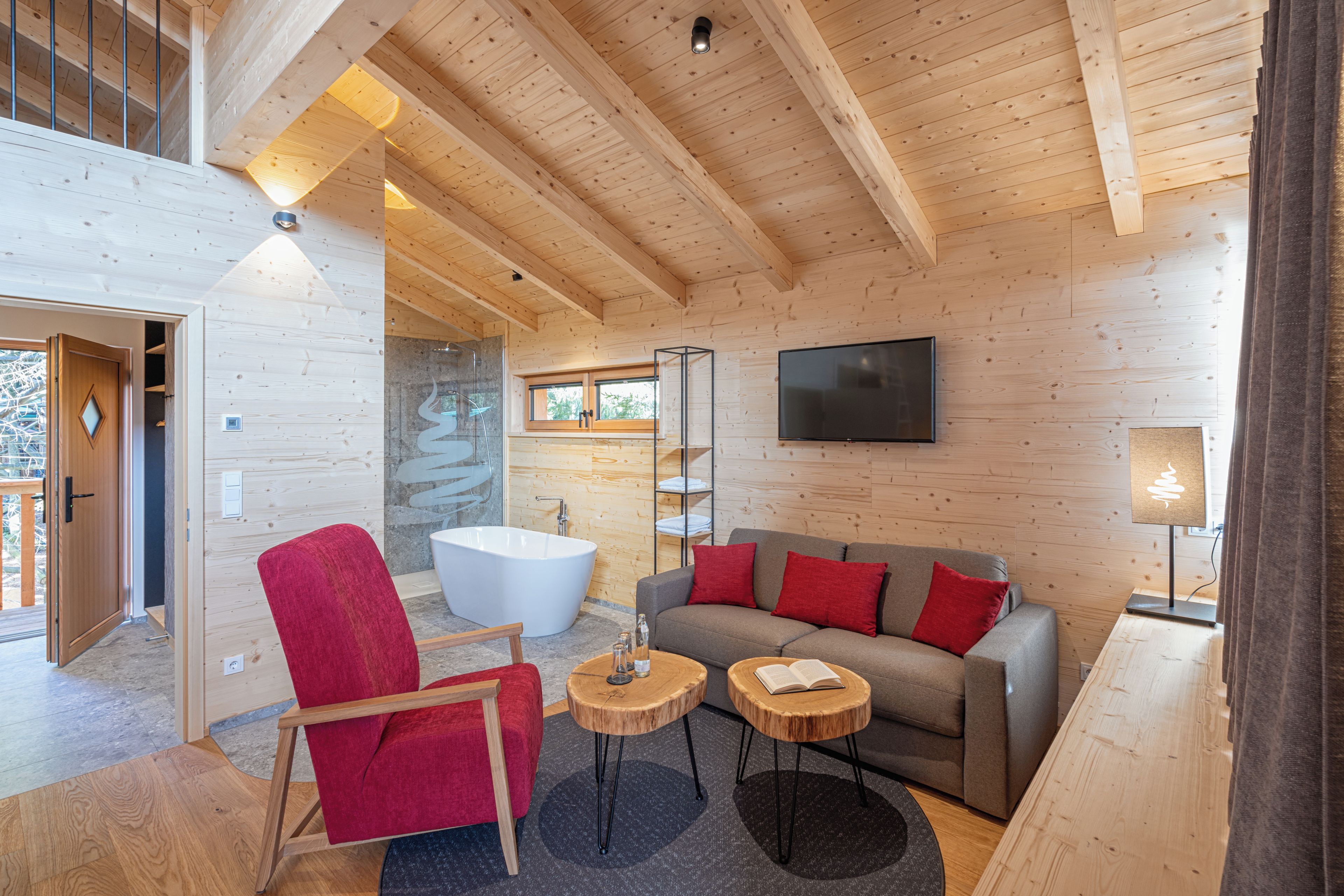 A modern interior with a wooden ceiling, a large bathtub, a grey sofa with red cushions, and a red chair.