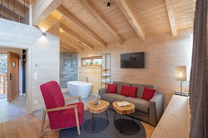 A modern interior with a wooden ceiling, a large bathtub, a grey sofa with red cushions, and a red chair. - Baumkronenweg GmbH (Kopfing im Innkreis)