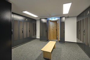 Un couloir avec des casiers, un banc en bois et une porte sur laquelle est apposée une signalisation verte indiquant la sortie.
