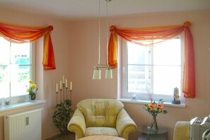 Ein Zimmer mit einem Sessel, einem Tisch, einem Heizkörper und einem Fenster mit Vorhängen.