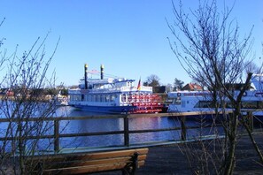 Ein Flussufer mit einem großen Boot, das an einem Steg festgemacht ist, einer hölzernen Bank und kahlen Bäumen.