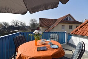 Un tavolo apparecchiato all'aperto, con una ringhiera blu, un libro, un vaso con fiori e un cielo sereno sullo sfondo.