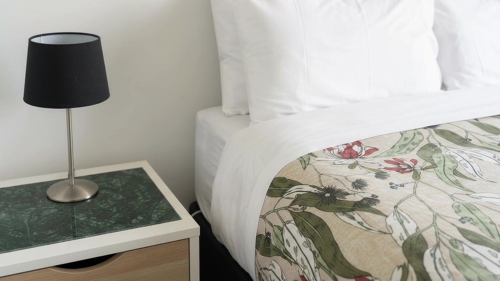 A bed with white bedding and a floral-patterned duvet, a bedside table with a black lamp, and a green marble-like surface.