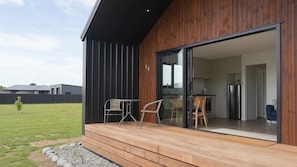 A modern house with a wooden deck, a sliding glass door leading to a kitchen, and a small outdoor seating area with two chairs. - Glaisnock Cottage Te Anau (Te Anau)
