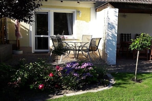 Eine Terrasse mit Tisch und Stühlen, ein kleiner Garten mit Blumen und ein Gebäude mit gelber Fassade.
