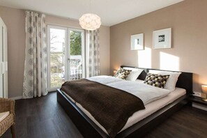 Un dormitorio con una cama grande, una ventana con cortinas, una mesita de noche con una lámpara y un cuadro colgado en la pared.
