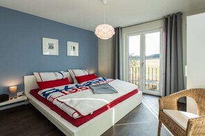 Ein modernes Schlafzimmer mit einem großen Bett, Blick ins Freie und einem Korbsessel.