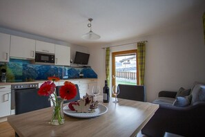 Eine moderne Küche mit weißen Schränken, einer Mikrowelle und einem Fernseher. Es steht ein Esstisch mit Blumen und einer Weinflasche darauf. Im Hintergrund ist eine Couch zu sehen.