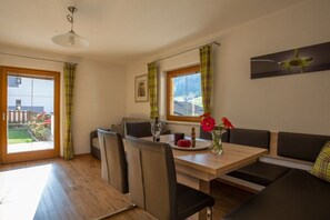Un coin repas avec une table en bois, des chaises et un banc. Il y a une fenêtre avec des rideaux, un ventilateur de plafond et un tableau encadré au mur.