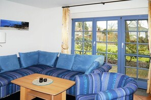 A room with a blue-striped sofa, a wooden table with two cups, and a large window with a view of a garden. - Oak Park W 2.2 || Ostseebad Prerow (Ostseebad Prerow)
