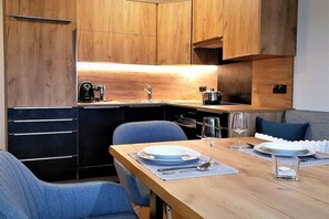 A compact kitchen with wooden cabinets, a built-in oven, and a dining area with blue chairs and a wooden table set for two. - at the Zedlacherhof guesthouse (Matrie in Osttirol)