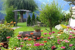 Ein Garten mit einer Holz-Pergola, verschiedenen Blumen und einer Bergkulisse.