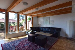 Ein geräumiges Wohnzimmer mit einem schwarzen Ledersofa, einem Couchtisch und einem gemusterten Teppich. Große Fenster lassen Tageslicht herein und bieten einen Ausblick ins Freie.