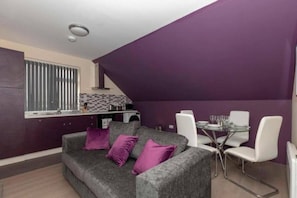 A compact living space with a kitchenette, a dining area with a glass table, and a grey sofa with purple cushions. - SAV Clarence Apt, Loughborough - 1-Bed Mandarin (Leicestershire)