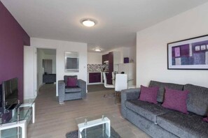 A modern living room with a grey sofa, a small armchair, and a glass coffee table. There is a television on the left and a dining area in the background. - SAV Clarence Apt, Loughborough - 2-Bed Charlies (Leicestershire)