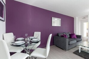 A modern living room with a dining area, featuring a purple wall, a grey sofa with cushions, and a glass coffee table. - SAV Clarence Apt, Loughborough - 2-Bed Charlies (Leicestershire)