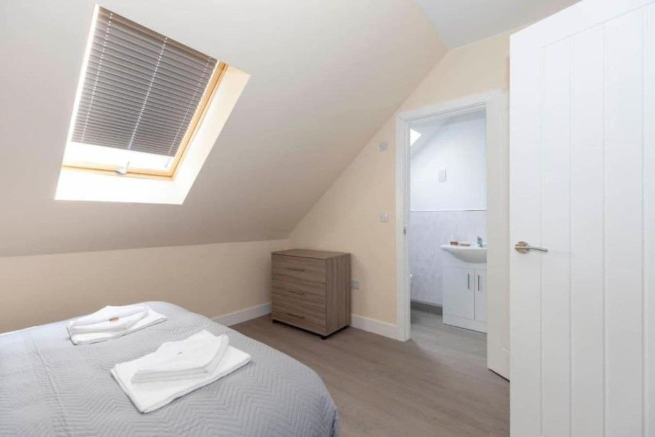 A bedroom with a sloped ceiling, a skylight, a bed with neatly folded white towels, a wooden dresser, and a door leading to a bathroom.