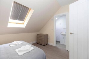 A bedroom with a sloped ceiling, a skylight, a bed with neatly folded white linens, a wooden dresser, and a white door leading to a bathroom. - SAV Clarence Apt, Loughborough - 1-Bed Beacon (Leicestershire)