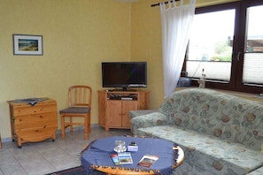 A living room with a television, a sofa, a chair, a small wooden cabinet, and a round table with books and a glass. - Holiday apartment with 1 bedroom (Wieck a. Darss)
