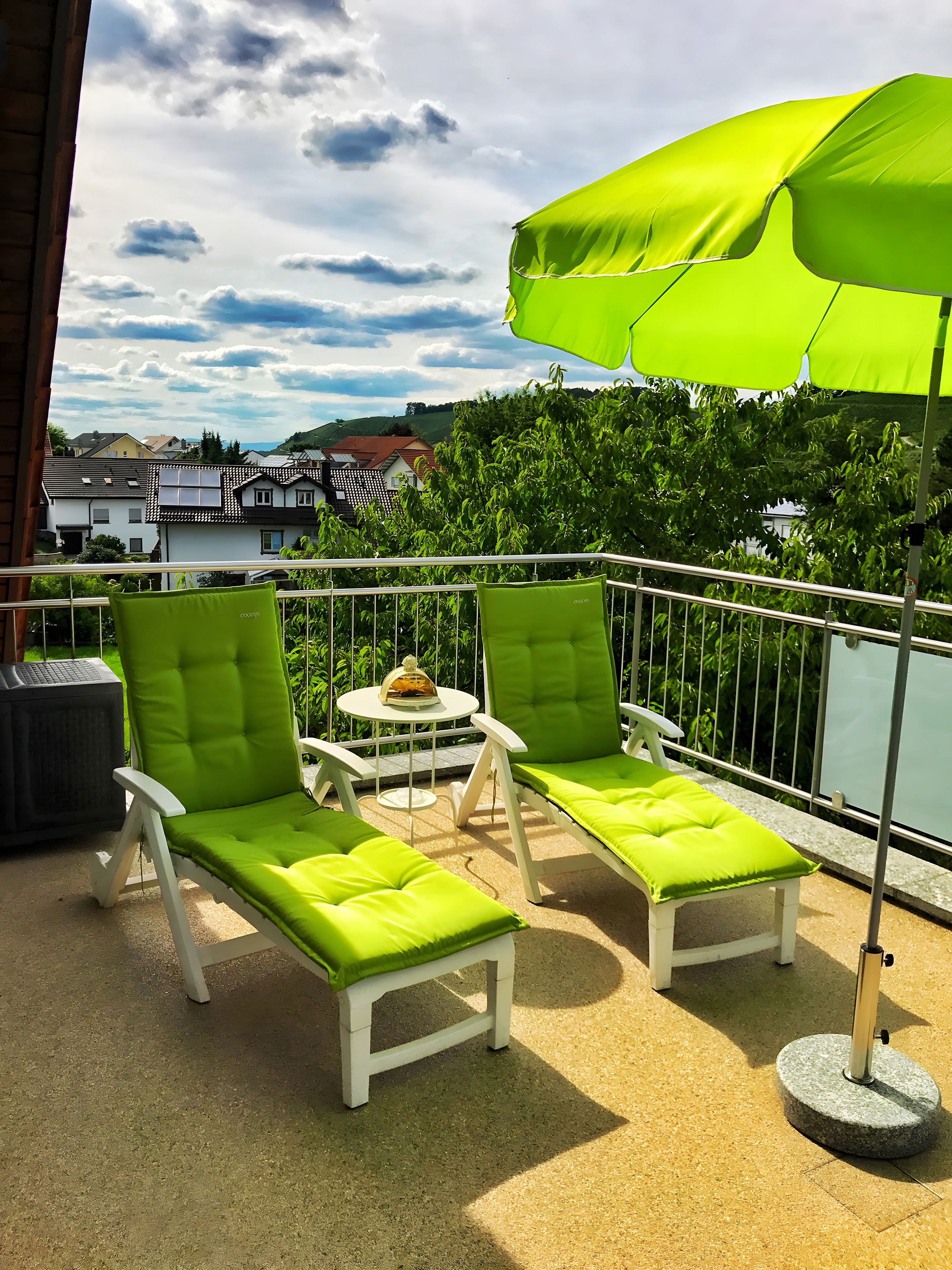 Een balkon met twee witte lounge fauteuils, een groene parasol en een klein, rond tafeltje met een decoratief object.