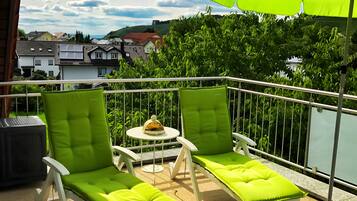 Een balkon met twee witte lounge fauteuils, een groene parasol en een klein, rond tafeltje met een decoratief object.