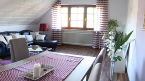 Una habitación con un sofá de cuero negro, una mesa de comedor con mantel a cuadros, un armario rojo y una ventana con cortinas a rayas.