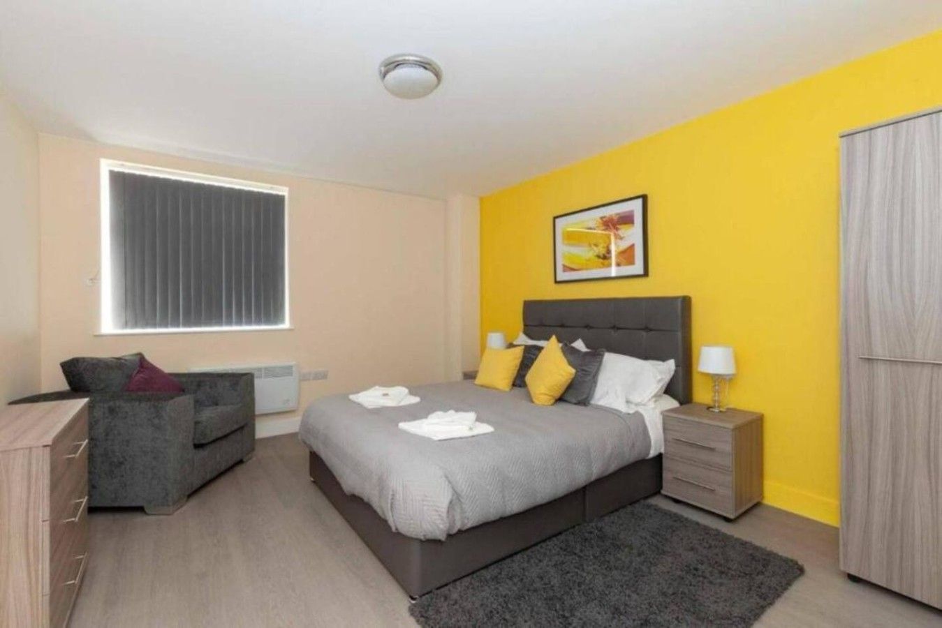 A bedroom with a bed, bedside tables, a dresser, a window with blinds, and a yellow accent wall.