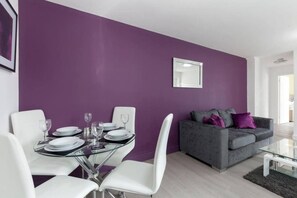 A modern living room with a dining area, featuring a purple wall, a grey sofa, and a glass coffee table. - SAV Clarence Apt, Loughborough - 2-Bed The Jack (Leicestershire)
