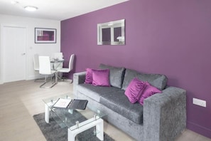 A living room with a grey sofa, two purple cushions, a glass coffee table, and a white dining area with chairs. - SAV Clarence Apt, Loughborough - 2-Bed Charnwood (Leicestershire)