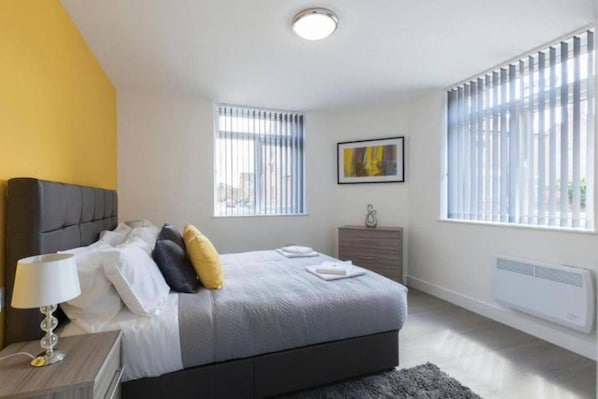A modern bedroom with a bed, bedside table, lamp, dresser, and windows. - SAV Clarence Apt, Loughborough - 2-Bed Charnwood (Leicestershire)