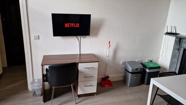 Une pièce comprenant un bureau, une chaise, un téléviseur fixé au mur, un balai, des poubelles et une petite table.