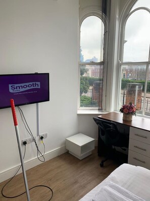 Une pièce avec un téléviseur diffusant la chaîne « Smooth Radio », un bureau avec une chaise, un petit meuble blanc et une fenêtre donnant sur des immeubles.
