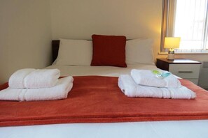 A hotel room with a bed, two towels, a red pillow, a nightstand, a lamp, and a window. - Sav Apartments Regent Leicester - Studio (Leicester)