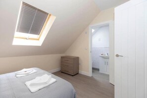 Ein Schlafzimmer mit geneigter Decke, einem Oberlicht, einem Bett mit ordentlich gefalteten weißen Handtüchern, einer Holzkommode und einer Tür zu einem Badezimmer.