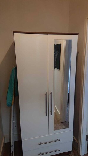 A white wardrobe with two doors and a mirrored section, located in a hallway with a towel hanging on the wall. - Sav Regent Leicester-Studio Walking to City Centre (Leicester)