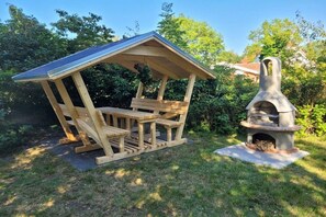 Eine hölzerne Laube mit geneigtem Dach und Bänken, neben einem Kamin aus Naturstein.