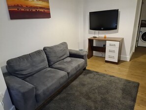 A grey sofa, a flat-screen TV, a wooden desk with drawers, and a washing machine in a room with a grey carpet. - SAV | Regent | Leicester-Studio Close to City Centre (Leicester)