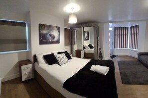 A modern hotel room with a large bed, a bedside table, a nightstand, a window with blinds, a painting on the wall, and a mirror. - SAV | Regent | Leicester-Studio Close to City Centre (Leicester)