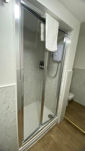 A glass-enclosed shower with a white towel hanging on the door, a handheld showerhead, and a toilet visible in the adjacent bathroom. - Sav | Regent | Leicester-Studio Walking to Demont (Leicester)