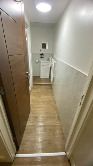 Un couloir étroit, doté d’un plancher en bois, mène à une cuisine avec des meubles blancs et une petite fenêtre.