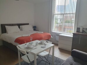 A bedroom with a bed, a dining table with chairs, a window with blinds, and a kitchenette area. - Sav Regent Leicester Twin Studio with Sofa bed (Leicester)