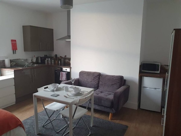 Ein kompakter Wohnraum mit Küchenzeile, einem Esstisch für zwei Personen, einem grauen Sofa und einem kleinen Kühlschrank.