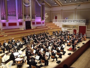 Ein großes Orchester in einer Konzerthalle mit abgestuften Sitzreihen und einem im Hintergrund sichtbaren, prägnanten Orgel.